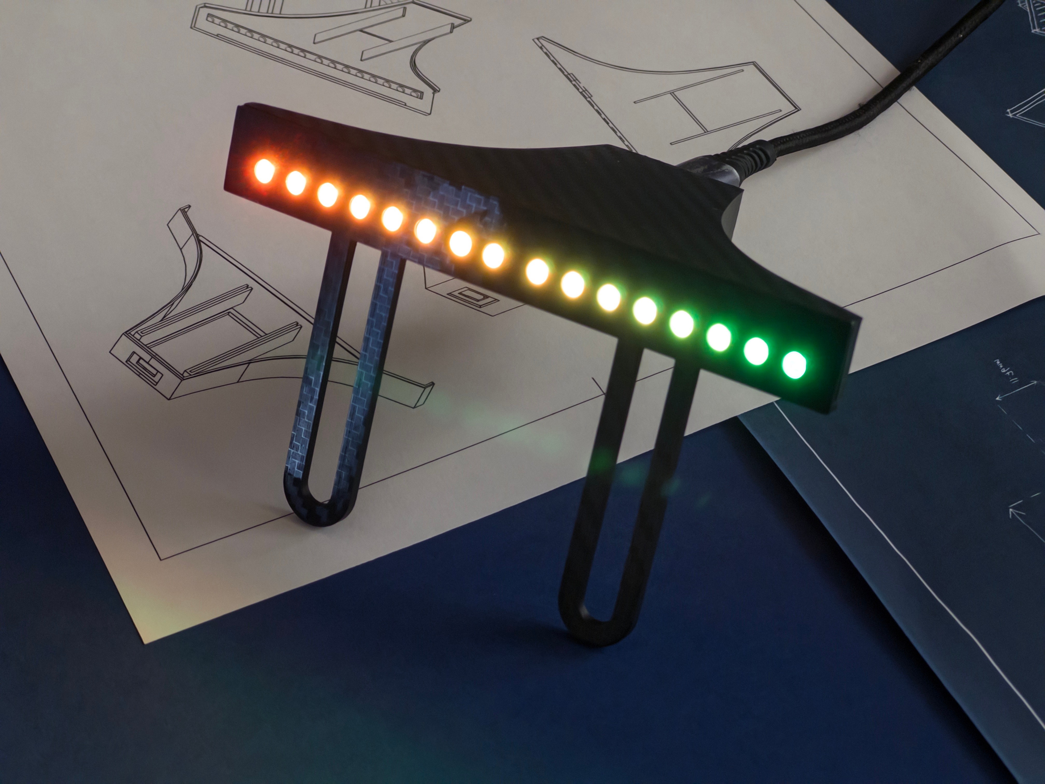 Rev Gauge sim racing shift light bar lit green to red across all 16 LEDs, carbon-fibre body on its Fanatec mounting legs, resting on design blueprints