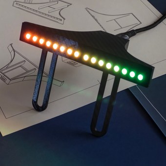 Rev Gauge sim racing shift light bar lit green to red across all 16 LEDs, carbon-fibre body on its Fanatec mounting legs, resting on design blueprints