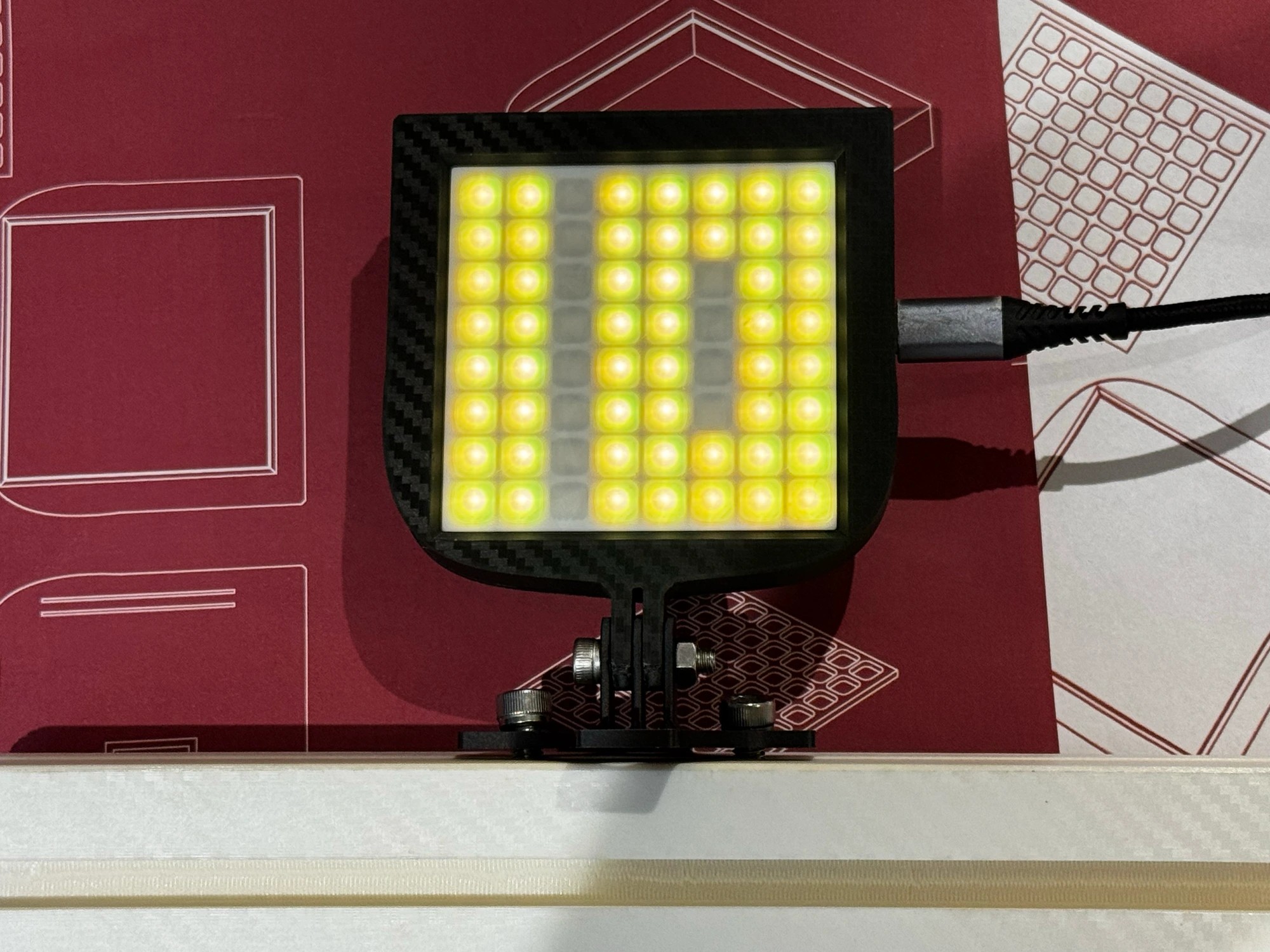 Close-up of the iFlag: GT Edition showing a bright yellow caution flag on the 8x8 RGB LED matrix in a carbon-fibre-effect housing, USB-C connected, on an oesimracing design blueprint