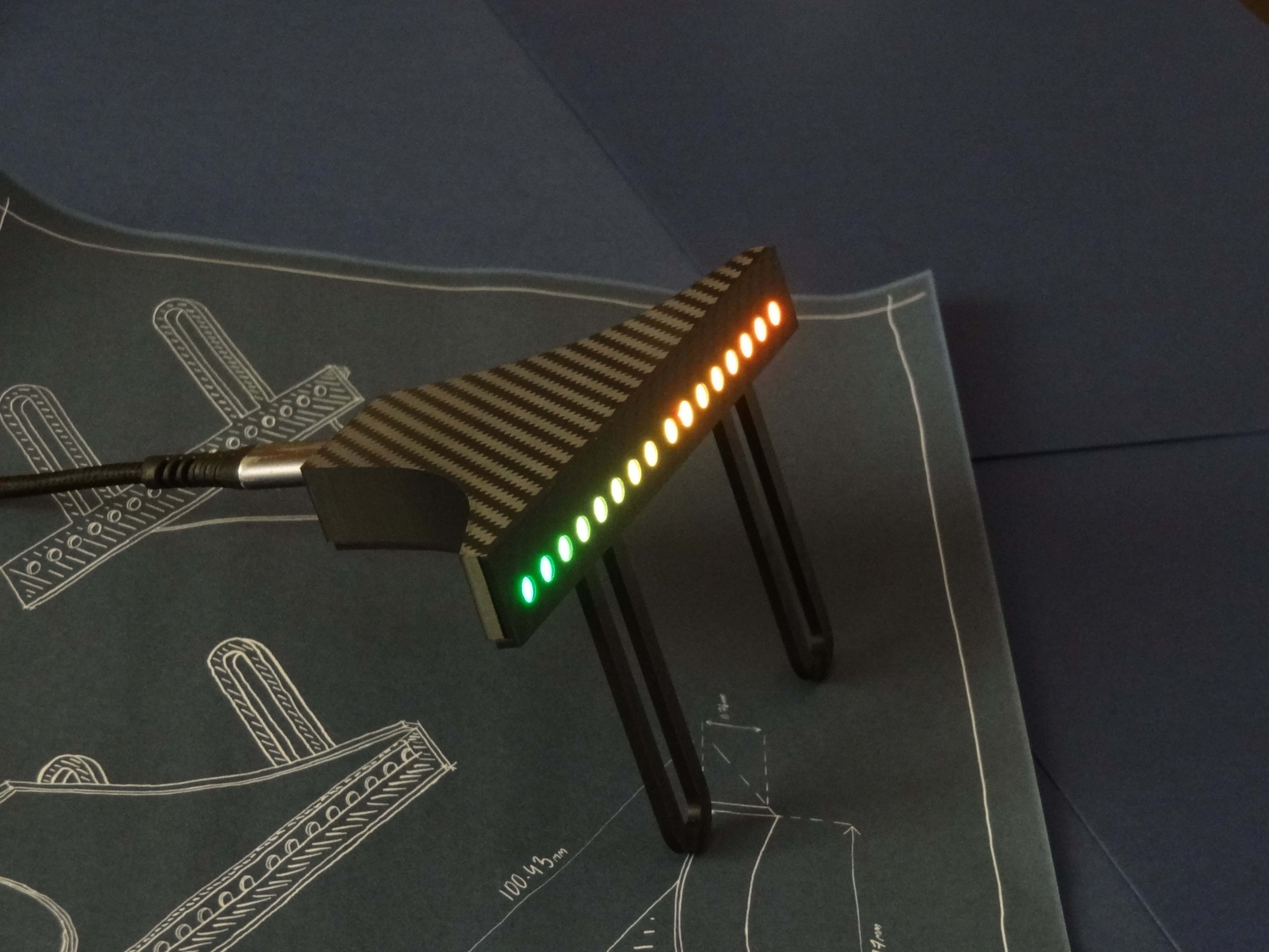 Angled close-up of the Rev Gauge shift light bar lit green to red, showing the carbon-fibre weave and USB-C connection